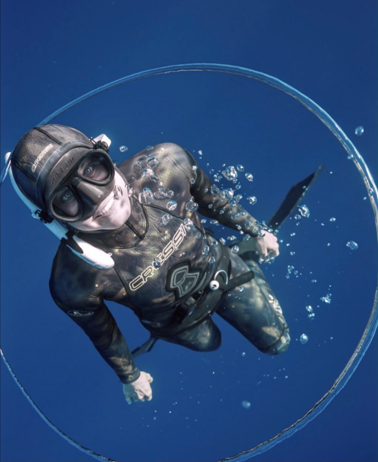 Tania Panés in a camouflage wetsuit and mask looking up through a ring of bubbles in clear blue water
