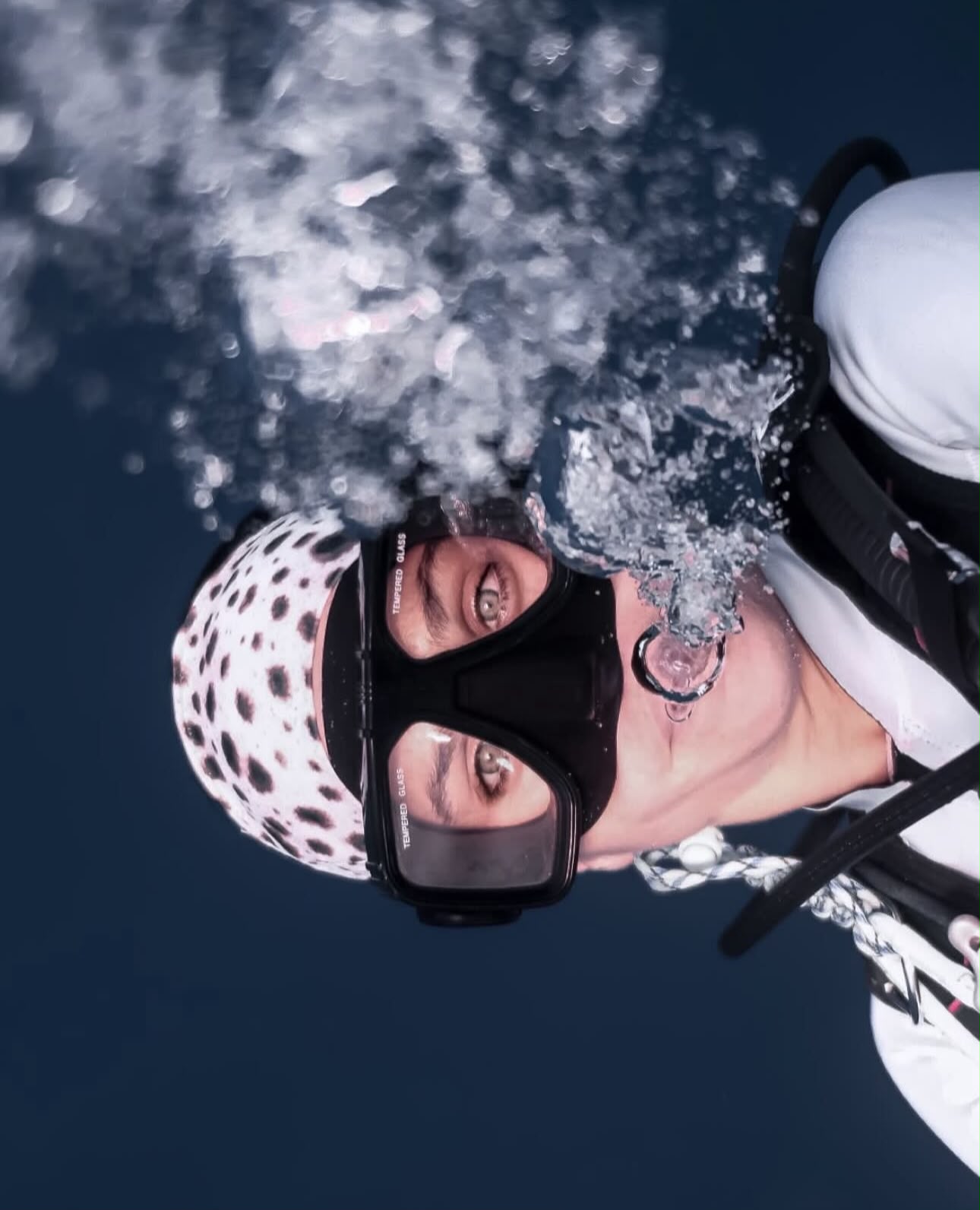 Tania Panés underwater in a head-down descent, looking at the camera through her dive mask while bubbles rise around her