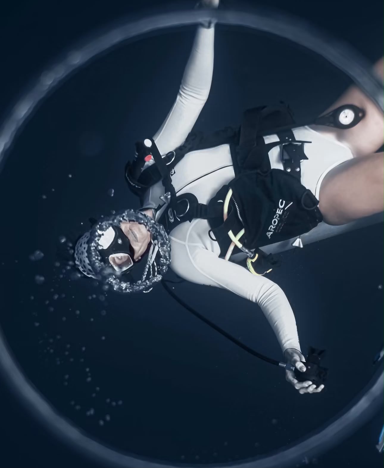 Tania Panés head-down in a white suit and black buoyancy harness, framed by a ring of bubbles against dark water